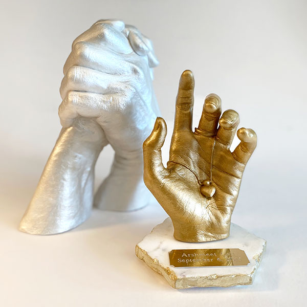 A gorgeous gold Individual Love Sculpture of a young woman's hand holding her necklace and locket, with an Opal Love Sculpture of a n older couple holding hands in the background